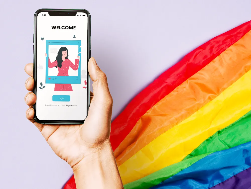 Hand holding a phone showing the app welcome screen in front of a rainbow flag